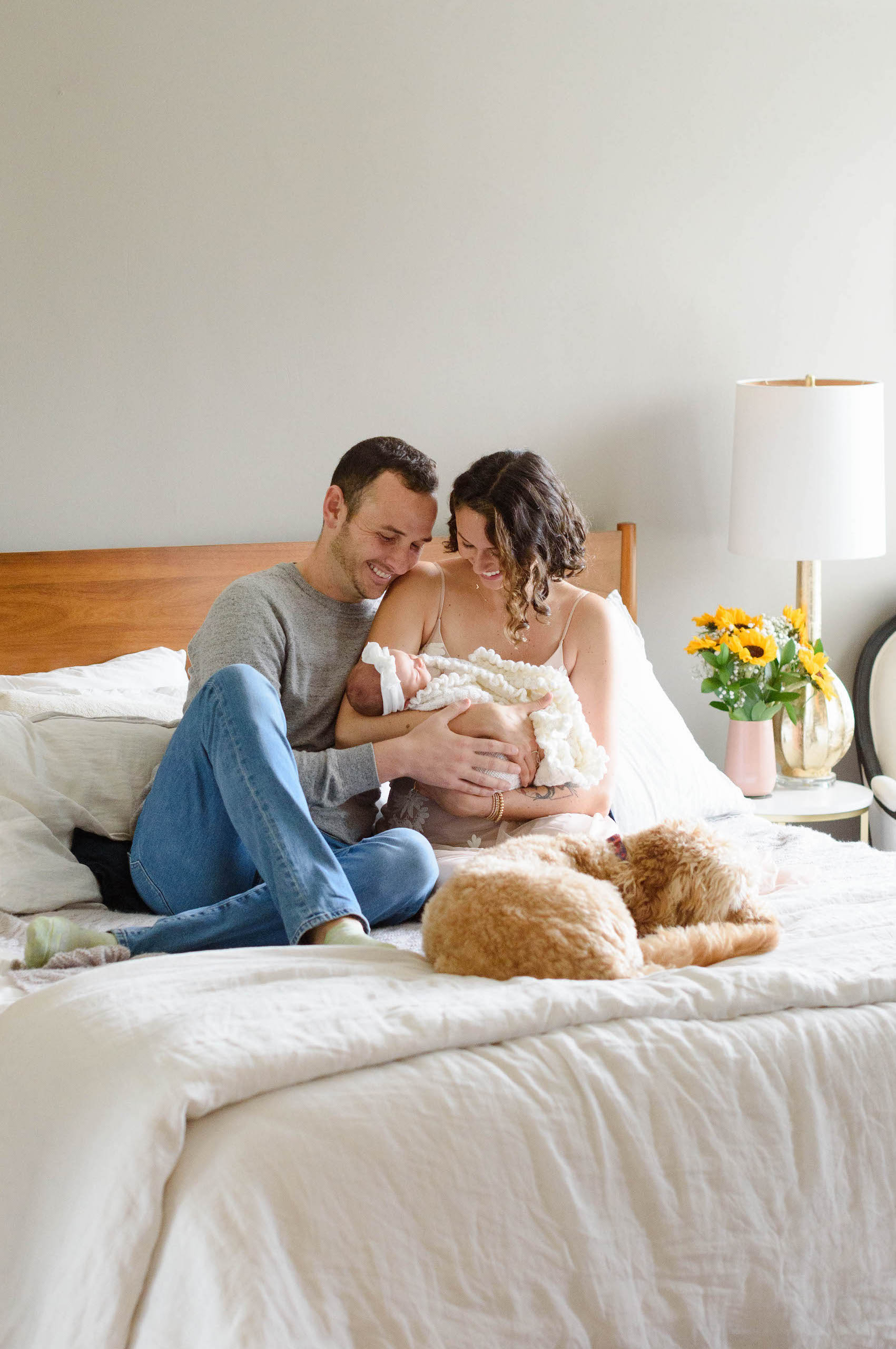 New parents sitting on bed looking at newborn baby with dog at their feet.